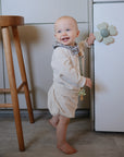 A smiling baby in a light outfit stands barefoot on the kitchen floor, holding a cabinet door with a flower knob. Beside the baby and stool are mushie Suction Spinner Toys, made from food-grade silicone for playful sensory fun.