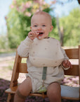 A smiling baby in a high chair outdoors wears a beige mushie Silicone Baby Bib and light clothes, holding food with greenery and pink blossoms softly blurred in the background.