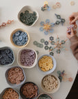 A child assembles colorful flower-shaped pieces from the mushie HEX Connect Building Toy Set on a white table with bowls of sorted pastel pieces, inspiring creativity and spatial reasoning.