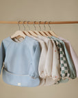 Six mushie Long Sleeve Bibs, featuring water-resistant fabric in pastel hues and playful patterns, hang on wooden hangers from a rod against a beige background.