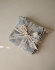 The mushie Muslin Cotton Washcloths, featuring a light blue and white checkered organic cotton design, are neatly folded and tied with a cream ribbon printed with the brand name, displayed on a neutral surface.
