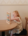 A young girl with Down syndrome sits at a round wooden table, focused on the Mushie Wooden Abacus. Dressed in a light floral dress, she perches on a dark cushioned chair in a softly lit room.