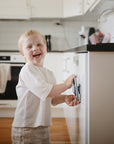 A young child smiles while playing with mushie Suction Spinner Toys on a kitchen appliance, enjoying sensory fun in a modern kitchen with white cabinets and wooden floors.