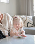 A smiling, light-haired baby in a bow and light outfit sits at a white table, holding the mushie Suction Spinner Toy made of food-grade silicone. An adult relaxes on a nearby sofa in the bright, cozy living room.