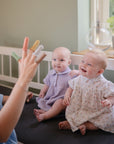 Two smiling babies sit on a dark mat as an adult displays mushie Silicone Animal Finger Puppets, encouraging fine motor skills. One baby wears a floral dress, the other lavender. A white crib and potted plant are in the background.