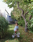 A toddler rides the mushie Mini Toddler Bike on a grassy path under a tree.