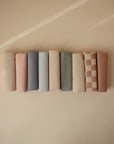 Nine neatly rolled Mushie Changing Pad Liners in soft, neutral solids—cream, grey, blue, green, brown—and a brown-and-cream checkered design are arranged against a beige background, highlighting the cozy organic cotton fabric.