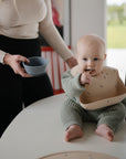 A baby in a green outfit sits on a table wearing a mushie Silicone Baby Bib and holding a spoon, while an adult stands nearby with a bowl in a bright, modern kitchen.