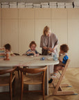 A woman serves food to three kids at a dining table, each wearing a mushie Silicone Baby Bib with an adjustable neck strap for easy clean-up. Toys, dishes, and an abacus are scattered across the cozy room’s table.