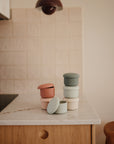 Four mushie Silicone Baby Food Containers in muted pink, green, and cream with ribbed lids are stacked on a marble countertop in a minimalist kitchen with tiled backsplash and wooden cabinets.