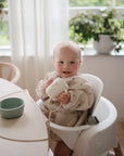 A smiling baby in a high chair holds a small cup, with baby essentials like a bowl and placemat on the table. The mushie Adjustable Silicone Strap 2-Pack keeps the pacifier nearby in the bright, airy room.