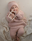 A smiling baby in pink lies on a cream blanket, holding the mushie No-Drop Sensory Teether. Subtle star patterns on the light background add charm to this adorable teething toy moment.