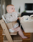 A baby in a striped outfit and bib sits in a wooden high chair at the table, with a mushie Adjustable Silicone Strap holding a whale-design sippy cup—one of the must-have baby essentials.