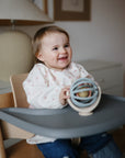A smiling toddler sits in a highchair, wearing the mushie Long Sleeve Bib with a subtle pattern, and holding a round, wooden and blue toy on the tray in a softly lit room.