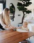 A woman with long blonde hair hands a green cup to a baby in a high chair wearing a mushie Silicone Baby Bib at a wooden dining table in a modern, bright living space with plants and neutral decor.