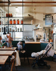 A woman sits at a cafe table with two young children wearing mushie Long Sleeve Bibs, while a baby sleeps in a stroller. The cozy, modern cafe has shelves of jars and a glass display case.