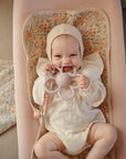 A smiling baby wearing a cream bonnet and outfit lies on a floral-patterned changing pad, holding the mushie Daisy Rattle Teether and looking at the camera.