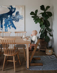 A baby sits in a high chair with the Mushie Splat Mat underfoot at a wooden dining table surrounded by chairs, a large plant, a blue abstract painting, and a woven basket in a bright, cozy dining room.