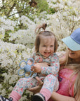 A smiling woman in a blue cap holds a laughing toddler in floral clothes among blooming white flowers. The toddler grips the mushie Daisy Rattle Teether, adding to the joyful moment.