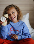 A smiling child in a blue "Buon Appetito" sweatshirt and red pants sits on a bed, holding mushie Suction Spinner Toys shaped like flowers and animals for a colorful sensory play experience.