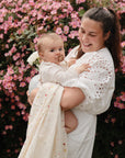 A woman in a white dress smiles while holding a baby with a pacifier before pink flowering bushes. The baby is wrapped snugly in the mushie Organic Cotton Muslin Swaddle Blanket.
