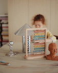 A child with curly hair sits at a table with the Mushie Wooden Abacus, a metallic lamp, a small sculpture, a clay bust, a wooden spoon, and a stack of books in the background.