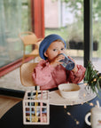 A young child in a blue knit hat and pink shirt drinks from a mushie Soft Spout Sippy Cup at a table with food, art supplies, and a cup, in a cozy modern room with large windows.