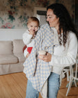 A woman, dressed in a white sweater and jeans, stands in a cozy living room holding her smiling baby—who wears red pants—and a blue-and-white checkered mushie Organic Cotton Muslin Swaddle Blanket along with a cup.
