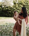A woman in a rust-colored dress holds a baby and the mushie Organic Cotton Muslin Swaddle Blanket outdoors, surrounded by greenery and scattered white flower petals on the grass.
