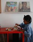 A young child in a blue shirt plays with mushie Wooden Balance Blocks on a red table, beside a vinyl record player and shelves filled with vinyl records, with two album covers displayed on the wall.