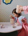 A young child in a pink dress plays at a table with the Mushie Silicone Tea Play Set, lifting the teapot lid while an adult pours from another teapot. The playful decor and wallpaper create a perfect setting for pretend tea parties.