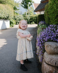 A young toddler in a light floral dress and black shoes stands on a path, smiling and holding a mushie Snack Cup, next to a stone wall with purple flowers and greenery under bright daylight.