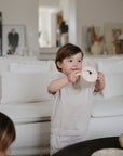 A young child smiles in a cozy living room with a white couch and framed photos, while another toddler holds a mushie Snack Cup—an easy-to-grip, spill-proof silicone cup perfect for snacks.