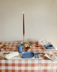 A table with a red-and-white checkered cloth is set with blue children’s dishes, two red candles, bread rolls, a mushie Silicone Baby Bib, and napkins tied with red string. Sunlight shines from the right.