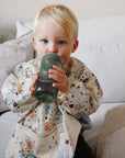 A young child with blonde hair sits on a couch, wearing a colorful space-themed bib and drinking from the mushie Soft Spout Sippy Cup in green, with neutral pillows and blankets in the background.