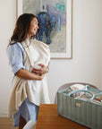 Lifestyle image of a woman holding a baby in a muslin nursing cover in a room with a Roman Green baby Diaper Caddy on a table.