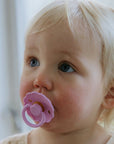 A young toddler with light blonde hair and blue eyes looks up while using a pink FRIGG Natural Rubber Pacifier from the FRIGG 4-Pack. Soft natural light illuminates their face, and a blurred indoor background is visible.