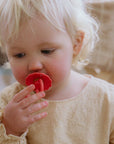 A young child with blonde hair thoughtfully looks down while holding a FRIGG Natural Rubber Pacifier from the FRIGG 4-Pack in their mouth, dressed in a light textured top against a soft, blurred background.