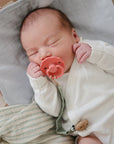 A newborn in a white onesie sleeps peacefully on a soft blanket, using a FRIGG Natural Rubber Pacifier from the FRIGG 4-Pack. Their eyes are closed, and the pacifier is gently held in their mouth.