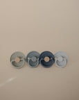 The FRIGG Natural Rubber Pacifier 4-Pack by FRIGG features four round pacifiers in muted gray, blue, and navy tones, neatly arranged against a neutral beige background.