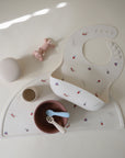 The mushie Silicone Baby Bib is shown with a matching bowl, cup, two spoons, and a silicone placemat in neutral tones on a light background, alongside pastel pink accessories. All items are easy to clean for everyday use.
