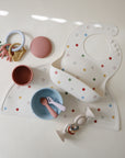 A flat lay of mushie feeding essentials: easy-to-clean Silicone Baby Bibs with adjustable neck straps, matching placemats, silicone bowls and utensils, a teether ring, pink cup, and pastel pacifier clips on a neutral background.