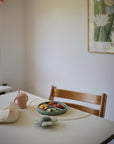 A small dining table features a mushie Suction Spinner Toy, a pink silicone sippy cup, and a plate of sliced vegetables and fruits. Soft lighting and minimalist decor with a “Tokyo” floral poster add to the gentle sensory ambiance.