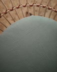 A close-up, top-down view of a round, cushioned seat with textured pale green fabric that resembles the mushie Mini Muslin Crib Sheet and features a curved, woven rattan or wicker backrest.