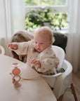 A baby with light curly hair sits in a high chair, reaching for a toy attached with a mushie Adjustable Silicone Strap 2-Pack. An adult is partially visible behind the baby. The background includes a window with sheer curtains and a green plant.