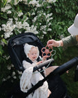 A baby in a stroller smiles while reaching for a flower-shaped toy attached to the mushie Adjustable Silicone Strap 2-Pack, held by a woman beside blooming white flowers and green bushes.