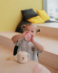 A young child with light hair in pigtails sits at a table, drinking from the mushie Soft Spout Sippy Cup. A mug and cookie are on the table, while bean bags and yellow cushions sit by the window behind her.