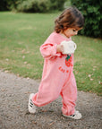Toddler in pink onesie walks outside holding a mushie Snack Cup in both hands.