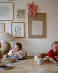 A woman and two kids laugh at a kitchen table covered in baking supplies, while the kids stay clean in mushie Long Sleeve Bibs. Framed art and a large bow decorate the wall behind them.