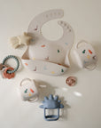 A flat lay of baby feeding essentials featuring the mushie Silicone Baby Bib with colorful dinosaur prints, two handled straw cups, a silicone teether ring with charms, a blue dinosaur teether, a star teether, and a cup lid.